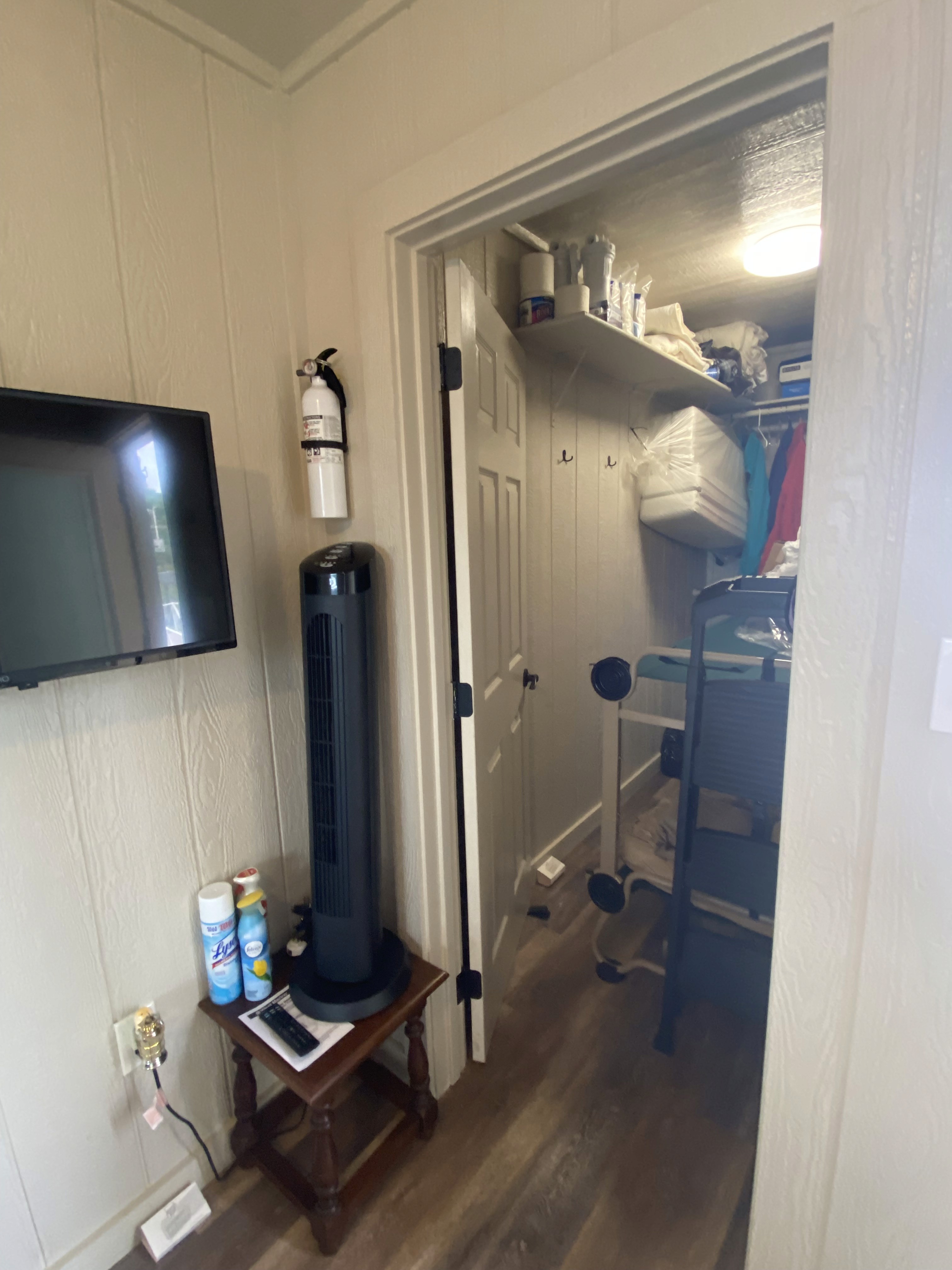 A narrow, well-organized closet in a home features neatly stacked towels and assorted supplies on shelves, with a folding step stool tucked away; a flat-screen TV and tower fan are seen on a wooden stand in a light-colored room.