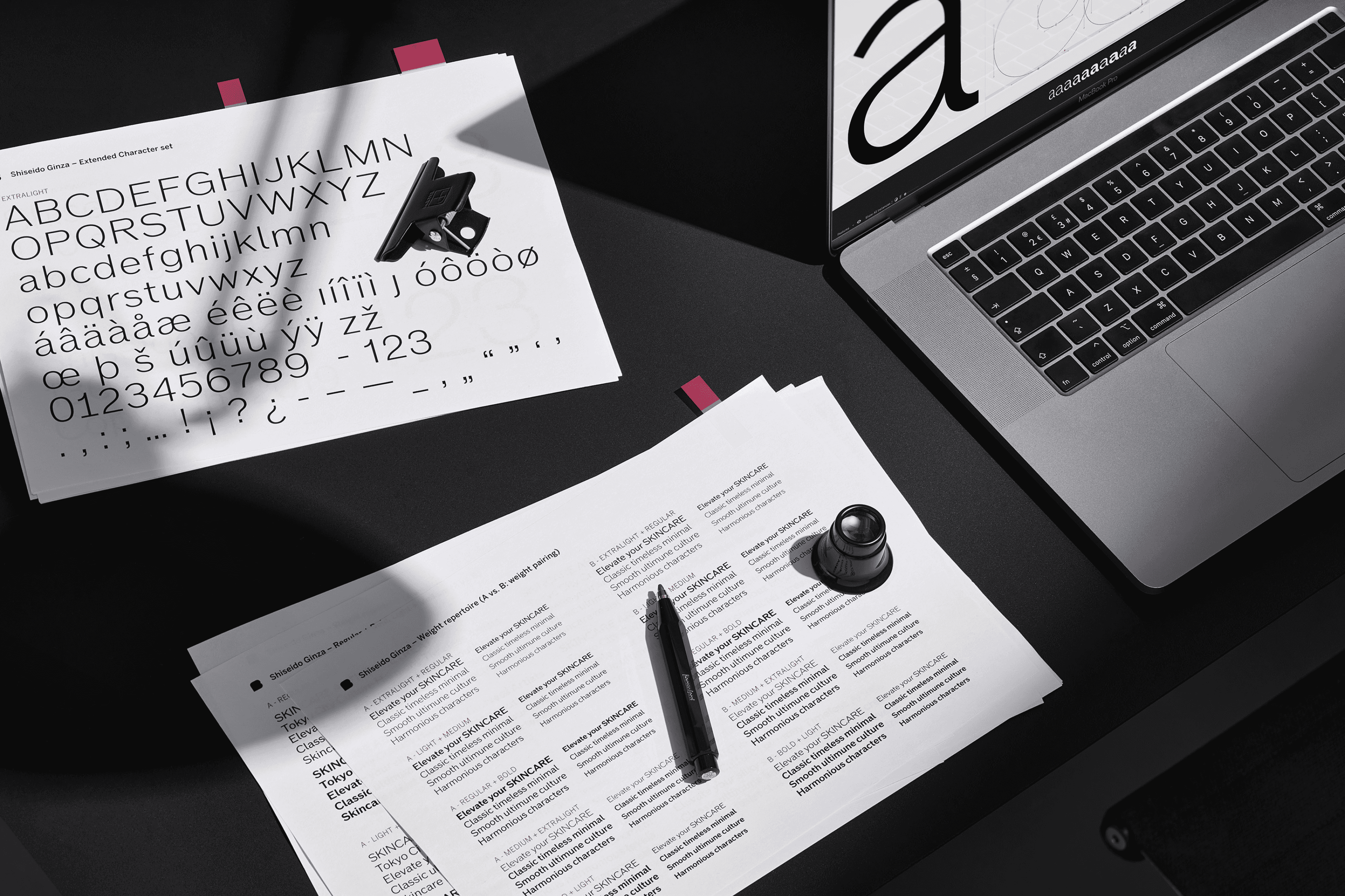 A hand holding a pen above papers with charts, next to a laptop on a dark surface.