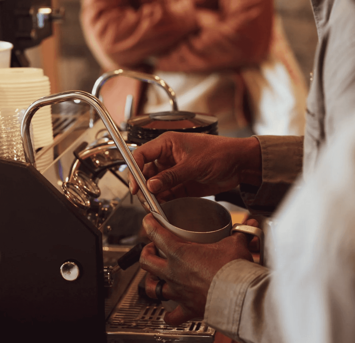 Close-up of a barista steaming milk with an espresso machine, showing hands holding a metal pitcher near the steam wand.