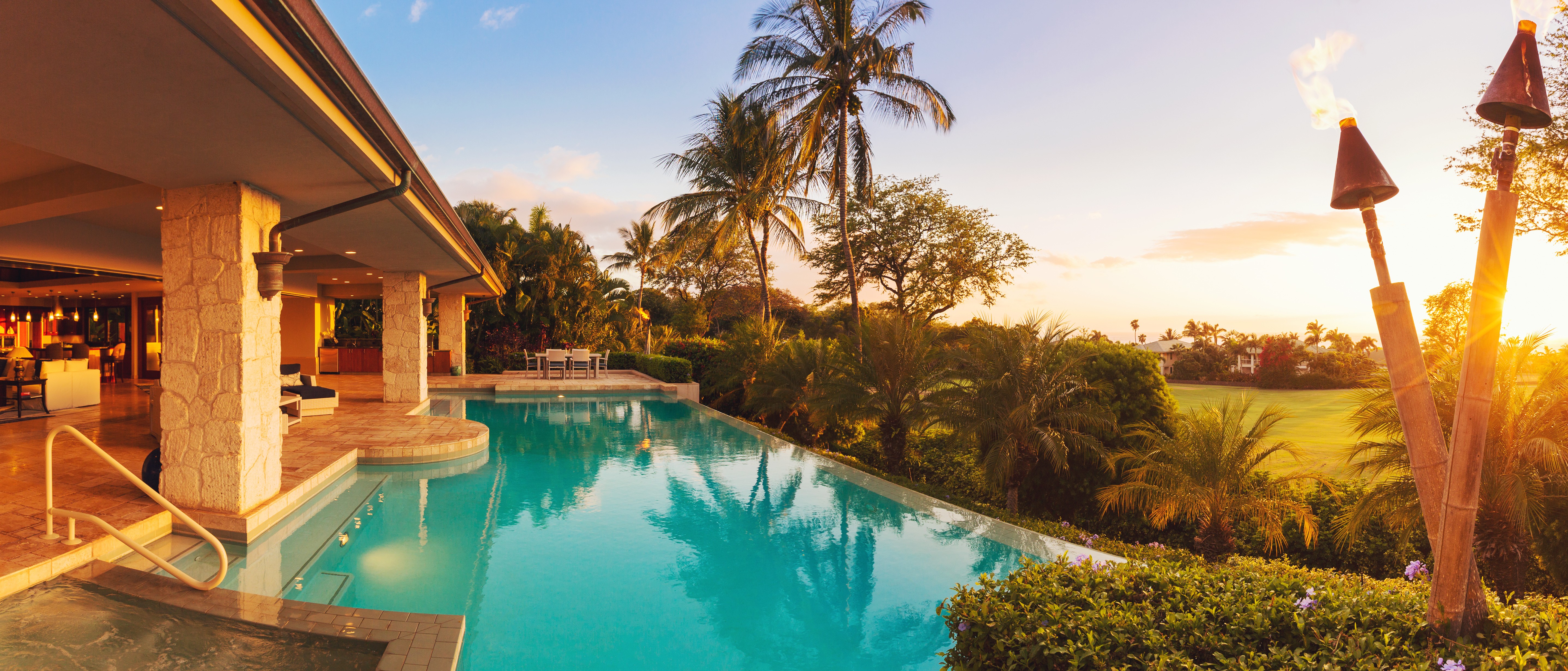Luxurious poolside scene at sunset with tropical palm trees, a serene infinity pool, and a spacious open patio. The sky glows warm with orange hues.