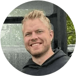 A smiling man with short brown hair in a dark blazer and white shirt against a light gray background. His expression conveys warmth and friendliness.