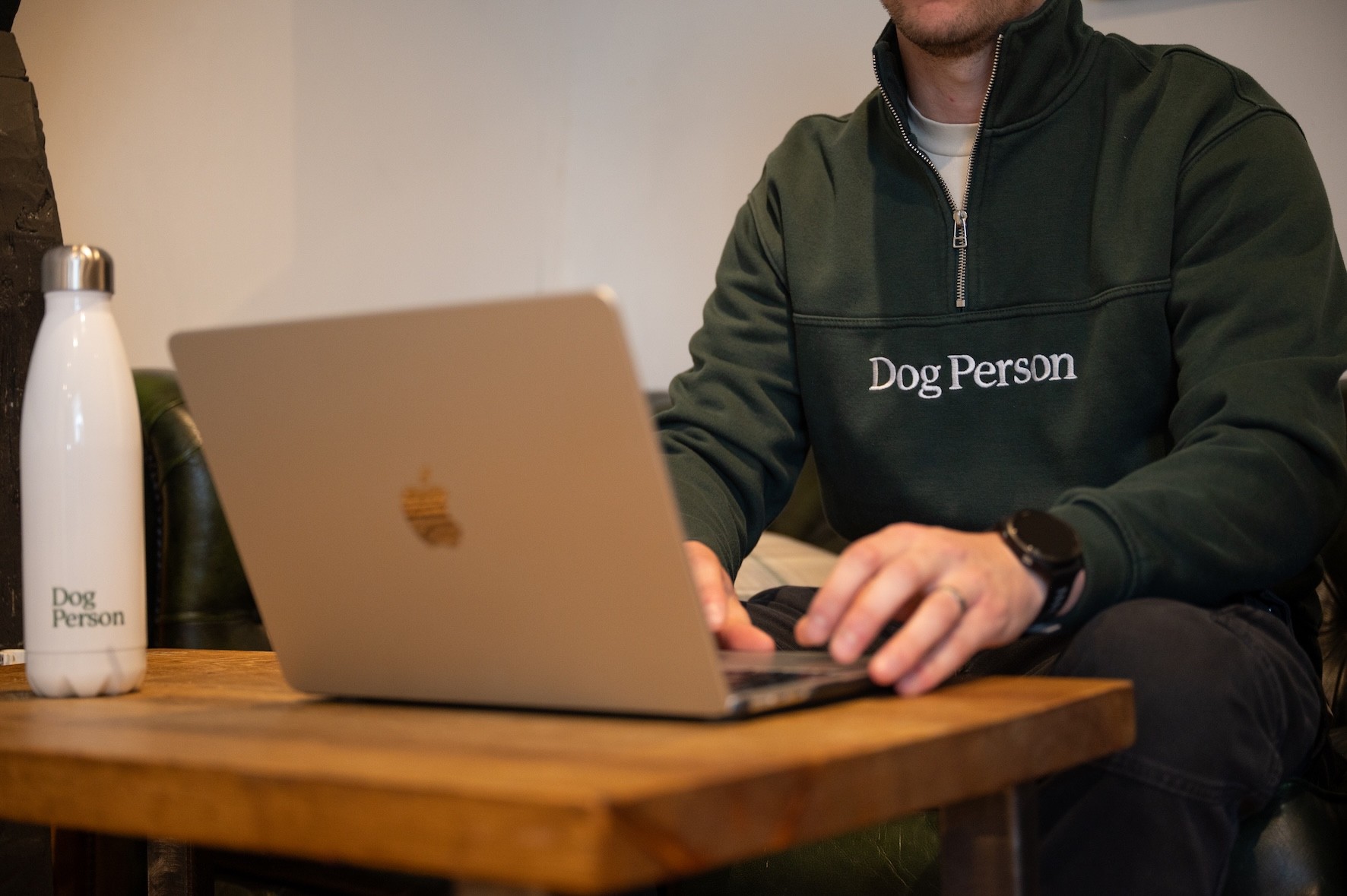 Scott (Dog Person) is sat at the laptop, with a Dog Person branded water bottle beside it. Scott is wearing a Dog Person branded jumper