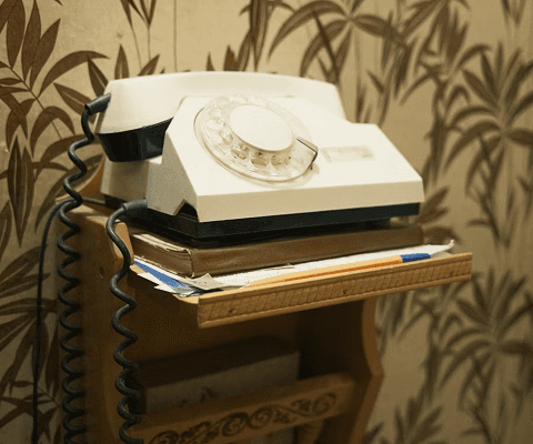 Vintage rotary telephone placed on a small wooden shelf, with papers stacked underneath and patterned wallpaper in the background.