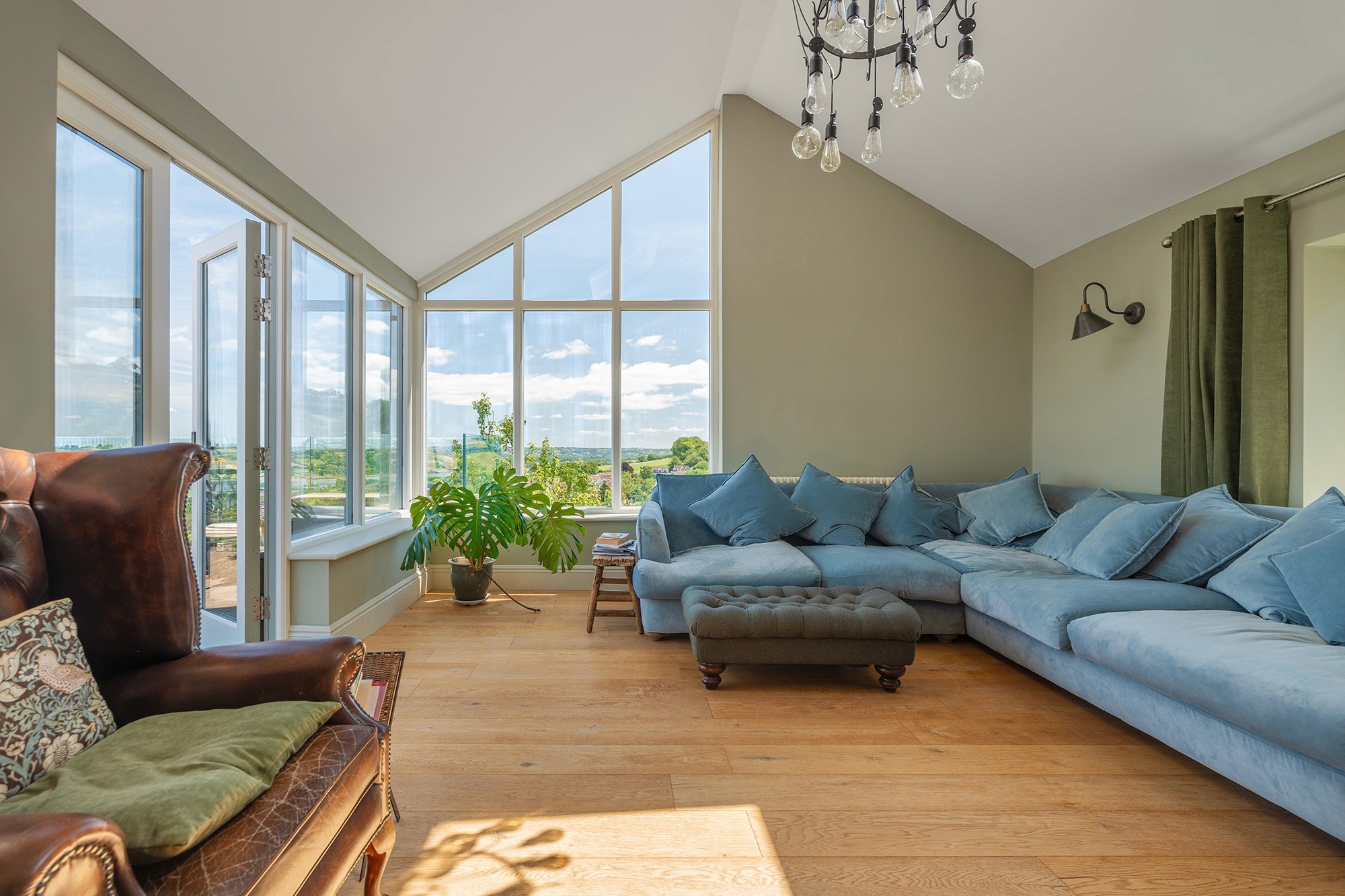 Bright, open-plan kitchen and dining area with a modern aesthetic. Features include soft green cabinetry, a grey stone accent wall, skylights, a glass table with a gold base, built-in upholstered seating, and a striking glass chandelier.