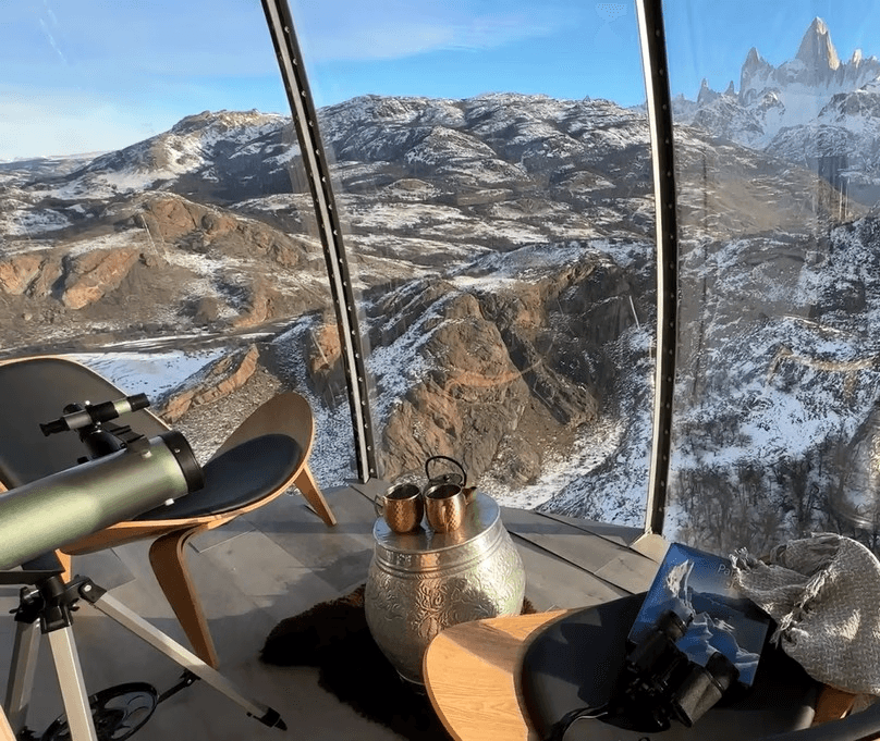 Vista desde cápsula acristalada: telescopio y binoculares junto a mesa metálica, ante montañas nevadas y el Fitz Roy al fondo.