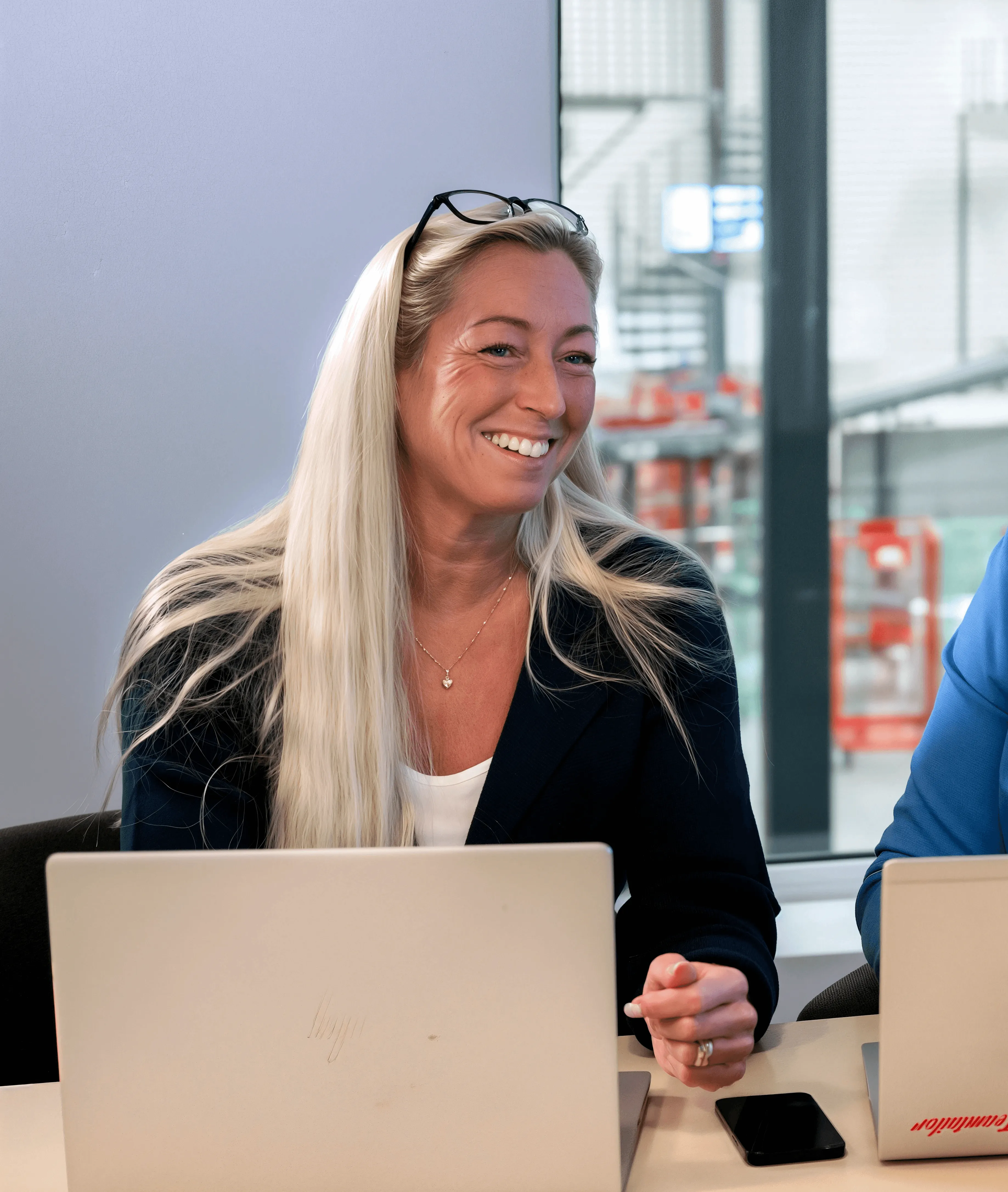Blond kvinna ler glatt vid bordet, bär glasögon, sitter framför en laptop.