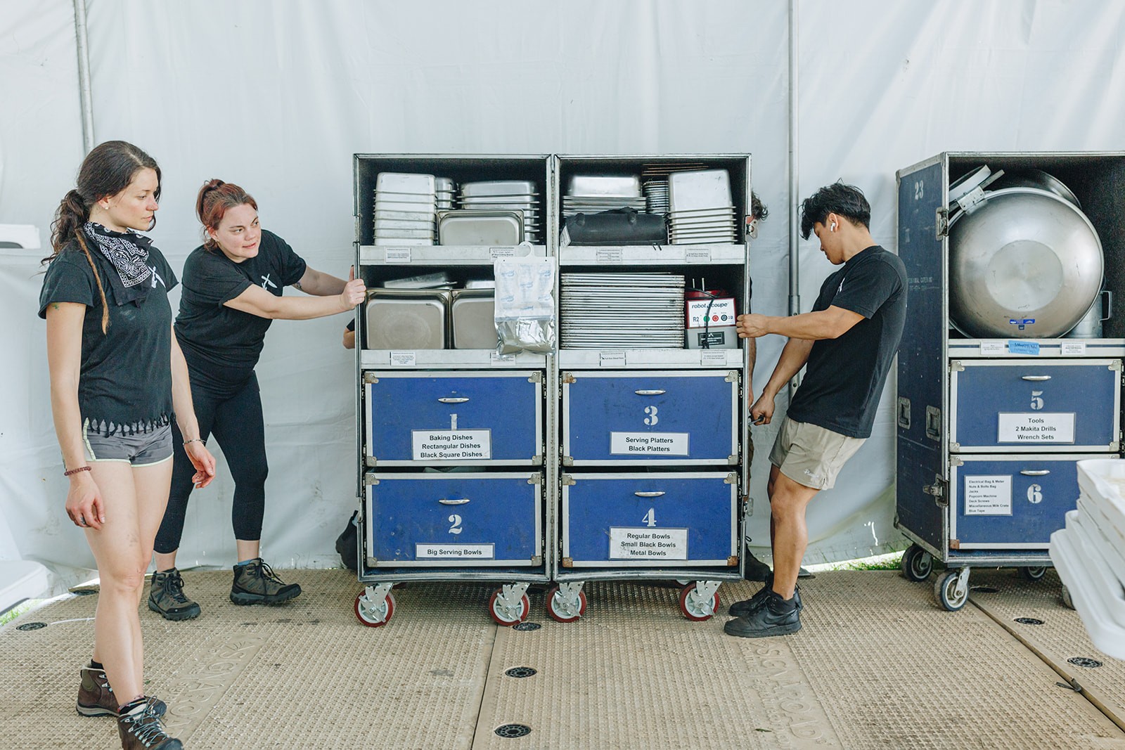 Two people organize equipment on carts in a large, white tent while another person stands nearby.