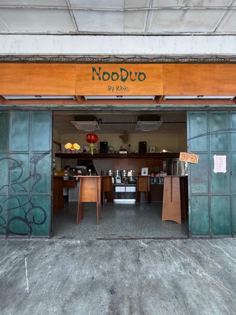 Exterior view of NoodDuo restaurant entrance with wooden sign, featuring 'By Kioku' below the main logo. The interior is partially visible with tables and decor, and there is a green metal door with a turtle graffiti design.