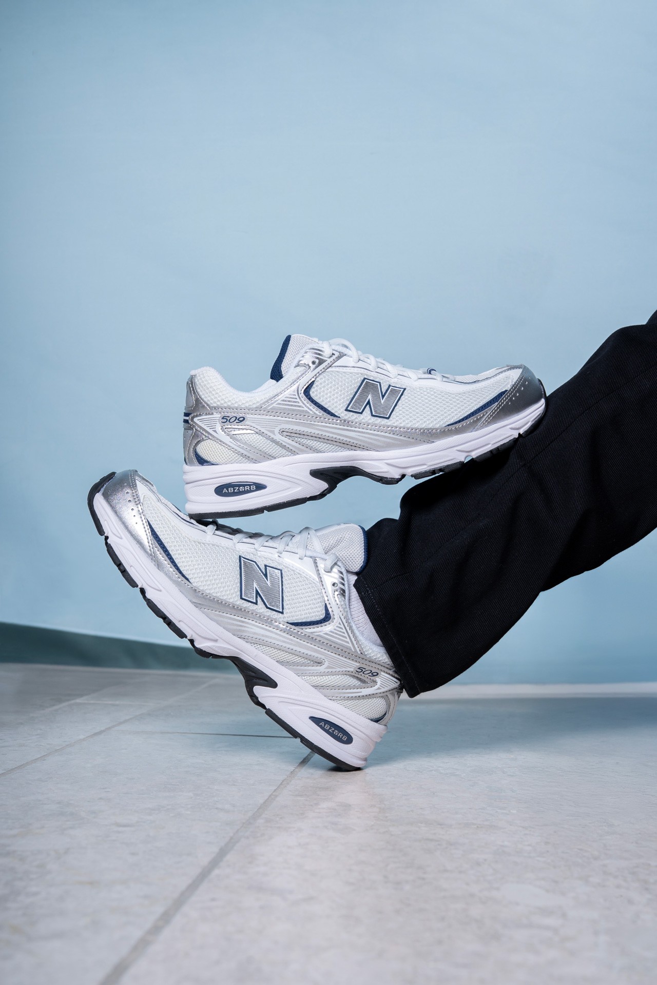 A styled close-up of white-and-silver New Balance 509 shoes — one on the foot, the other held against the lower leg, teal wall in the background.