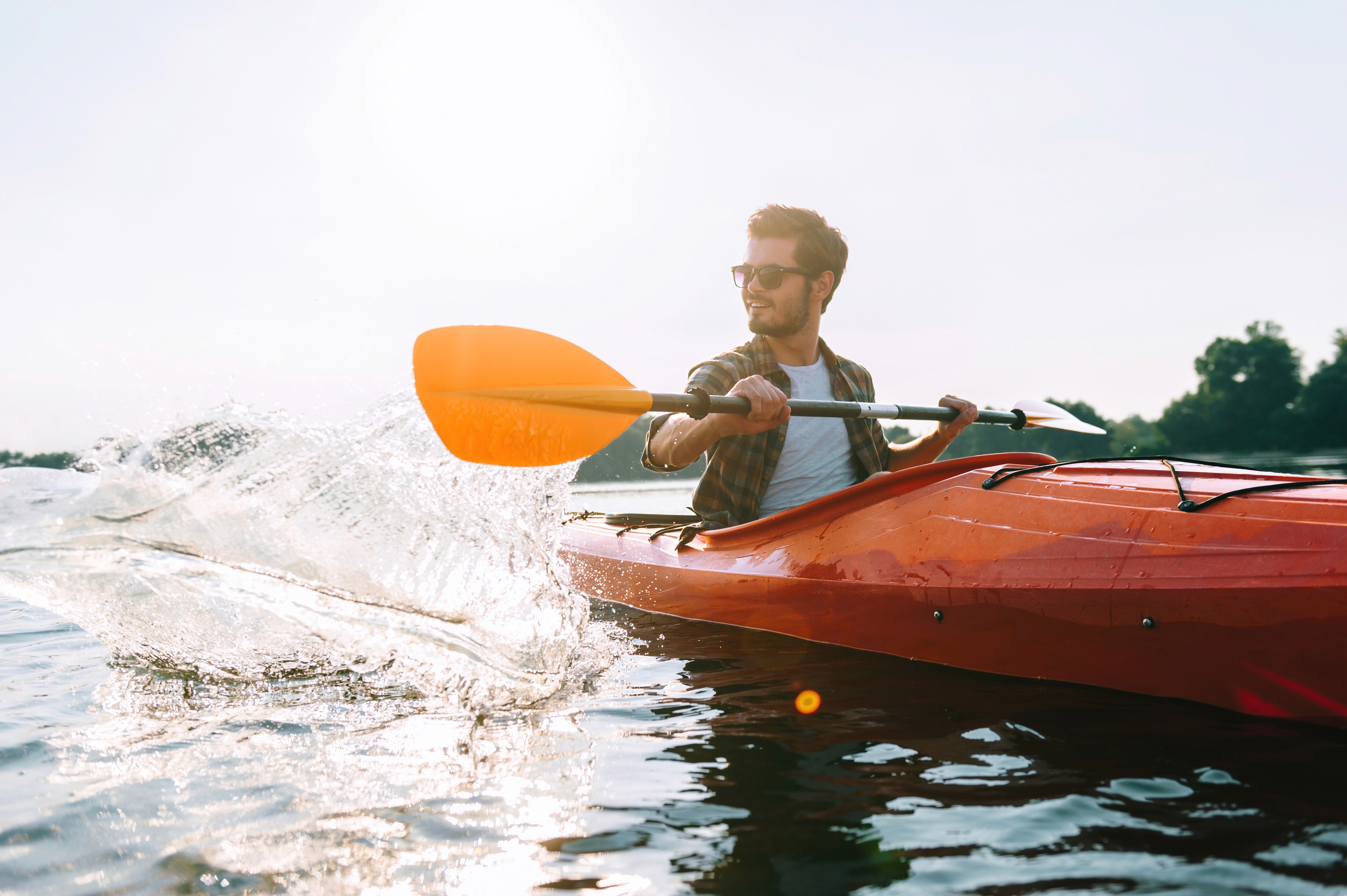 A person with dark hair in a ponytail, wearing a plaid shirt and a beige backpack, paddles a wooden canoe on a calm river