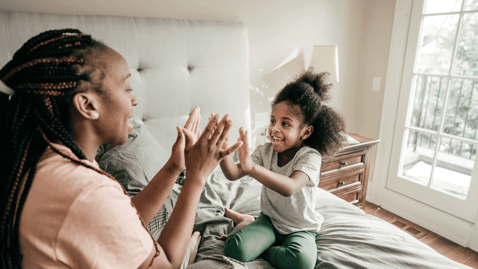 A mother and daughter sitting on a bed, pressing their hands together, smiling, and playing a game with each other.