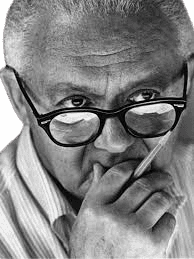 A black-and-white portrait of American graphic designer Paul Rand, known for his iconic corporate logo designs, including IBM and ABC. He is depicted wearing glasses, with a thoughtful expression, holding a pencil near his chin. The close-up image emphasizes Rand's intellectual and creative approach, reflecting his influence on modern graphic design and visual communication.