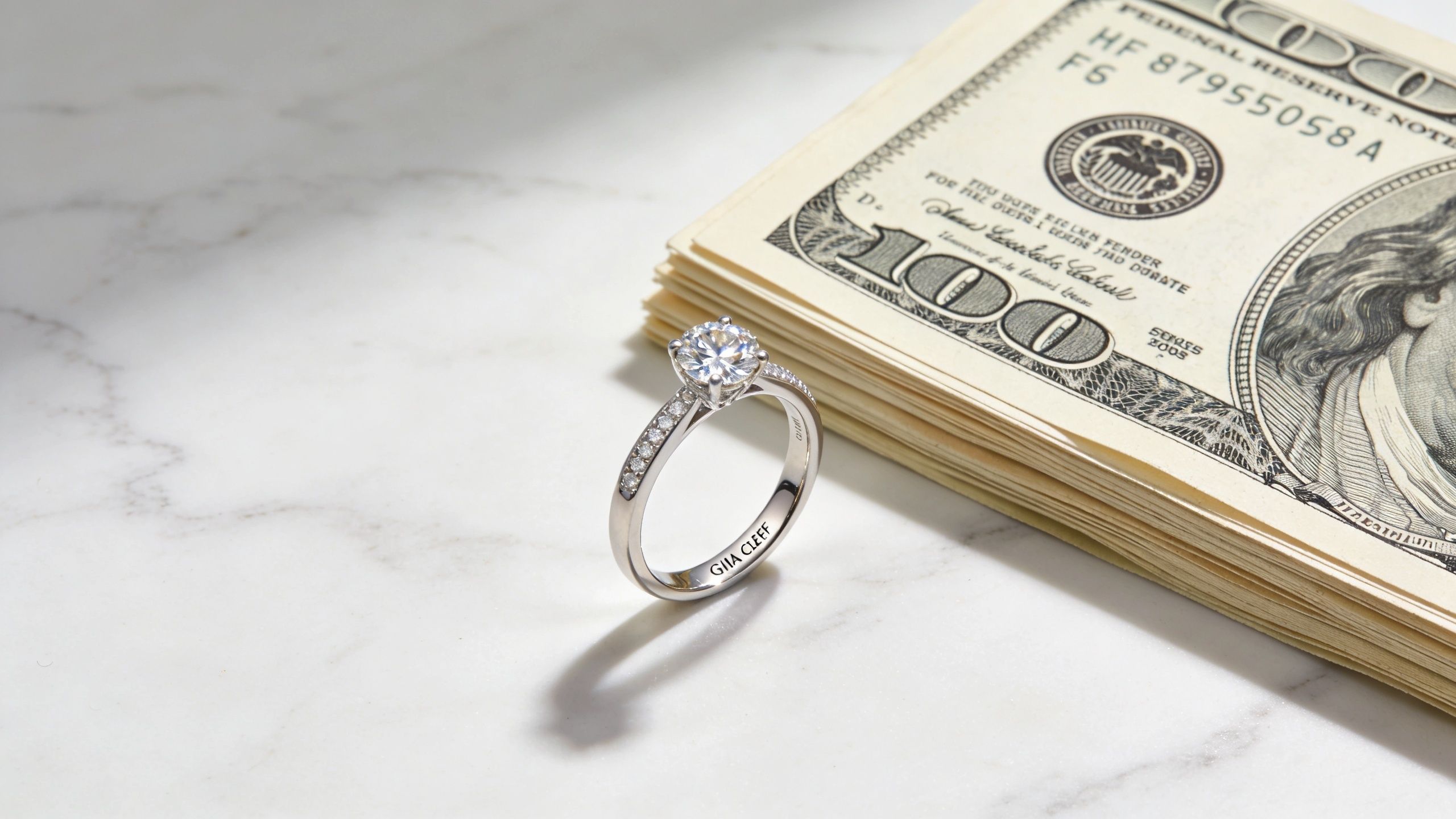A GIA Clef engagement ring with a round diamond center stone resting next to a stack of money.