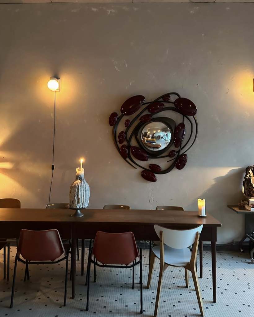 A cozy dining area with a long wooden table surrounded by various chairs. A decorative wall mirror encircled by red elements hangs above the table, complemented by a candle holder and warm lighting.