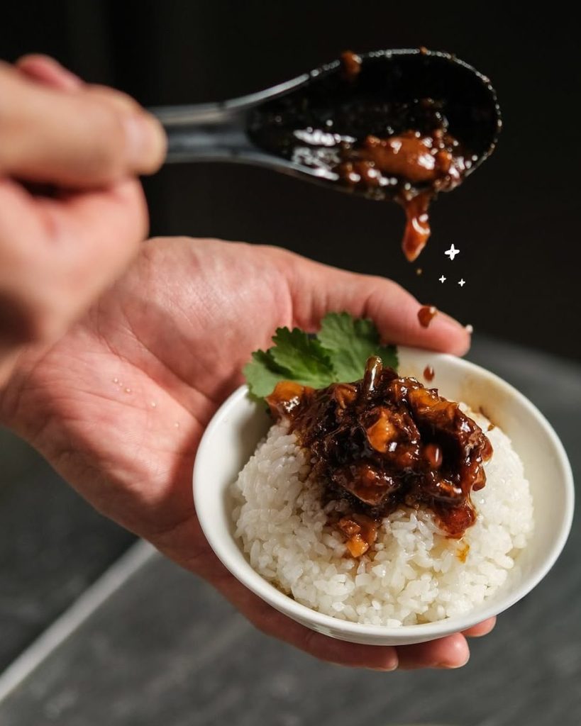A hand holding a bowl of rice topped with a rich, dark sauce and pieces of chicken, garnished with a sprig of cilantro.