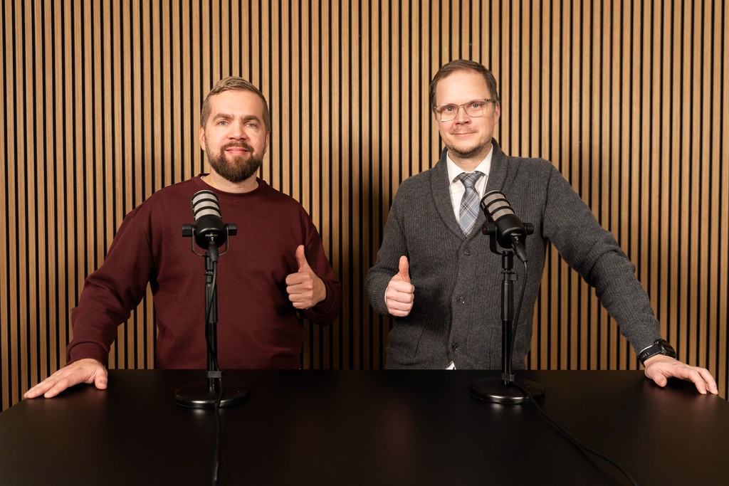 A smiling woman with her arms crossed, standing against a dark green background. She has long, dark hair.