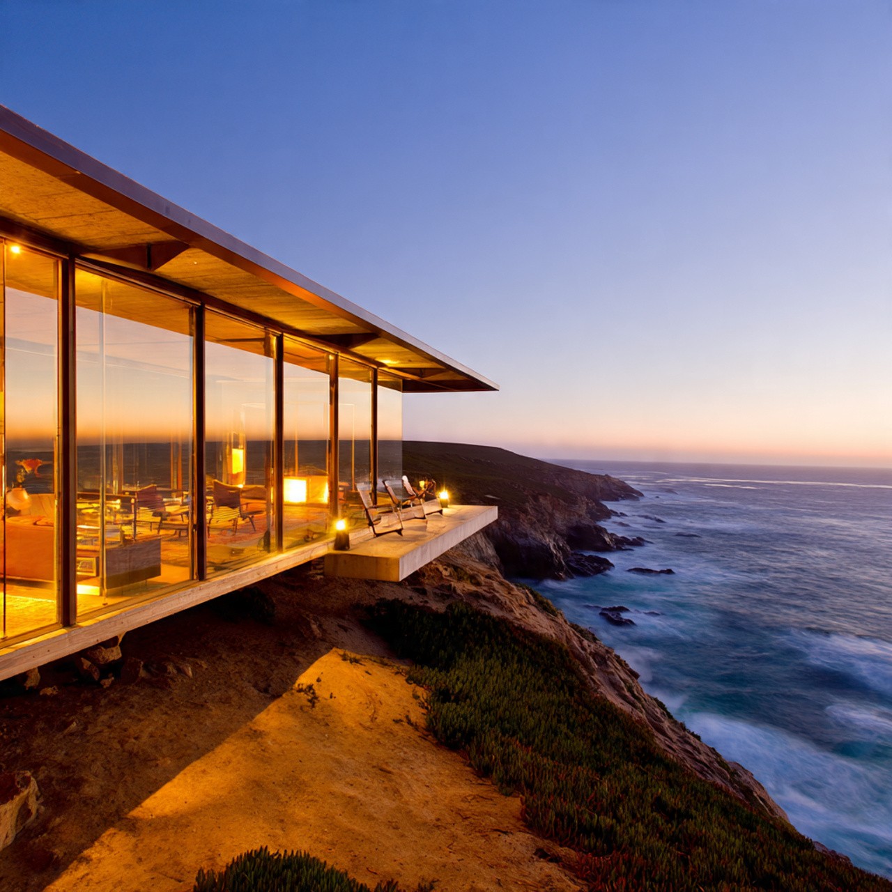 Modern glass house perched on a cliff at sunset, overlooking the ocean. Warm interior lights create a cozy contrast against the vast, serene seascape.