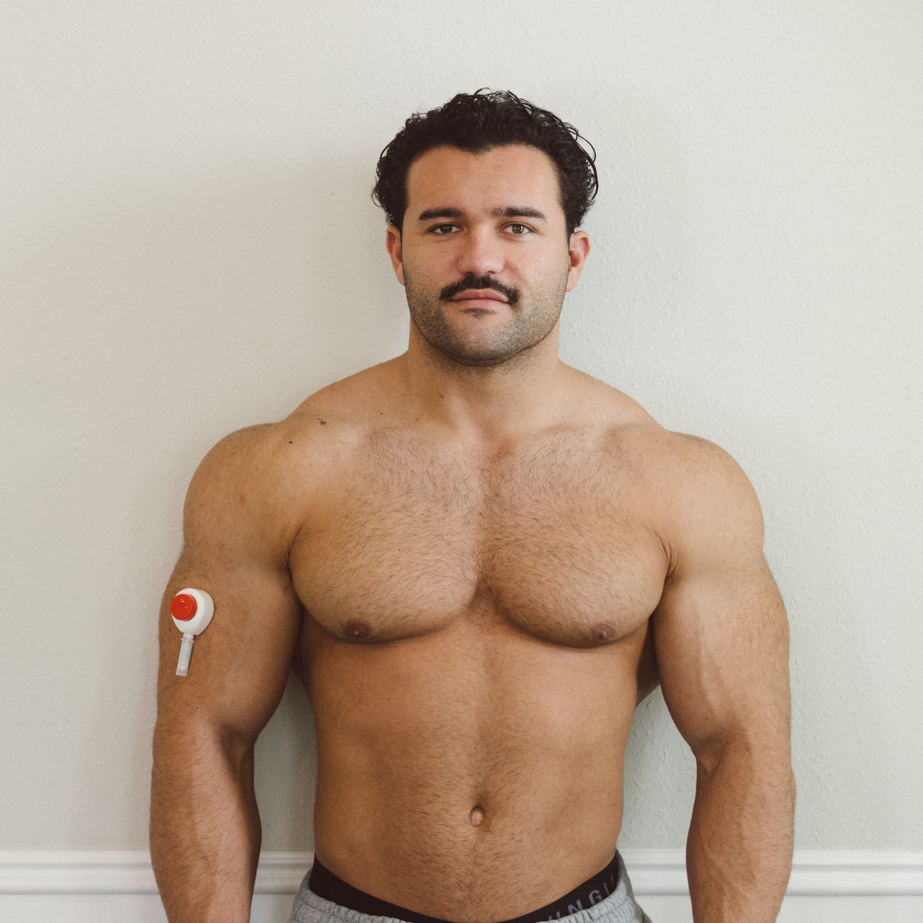 Muscular man standing against a neutral background wearing a Rythm Health wearable biosensor patch on his upper arm for at-home health and biomarker monitoring.