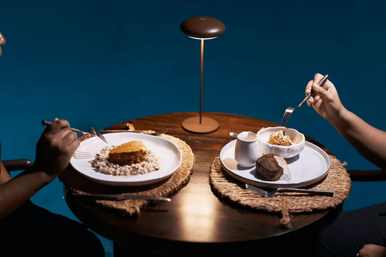 Two hands reaching for plates of food on a wooden table, with a dark blue background.