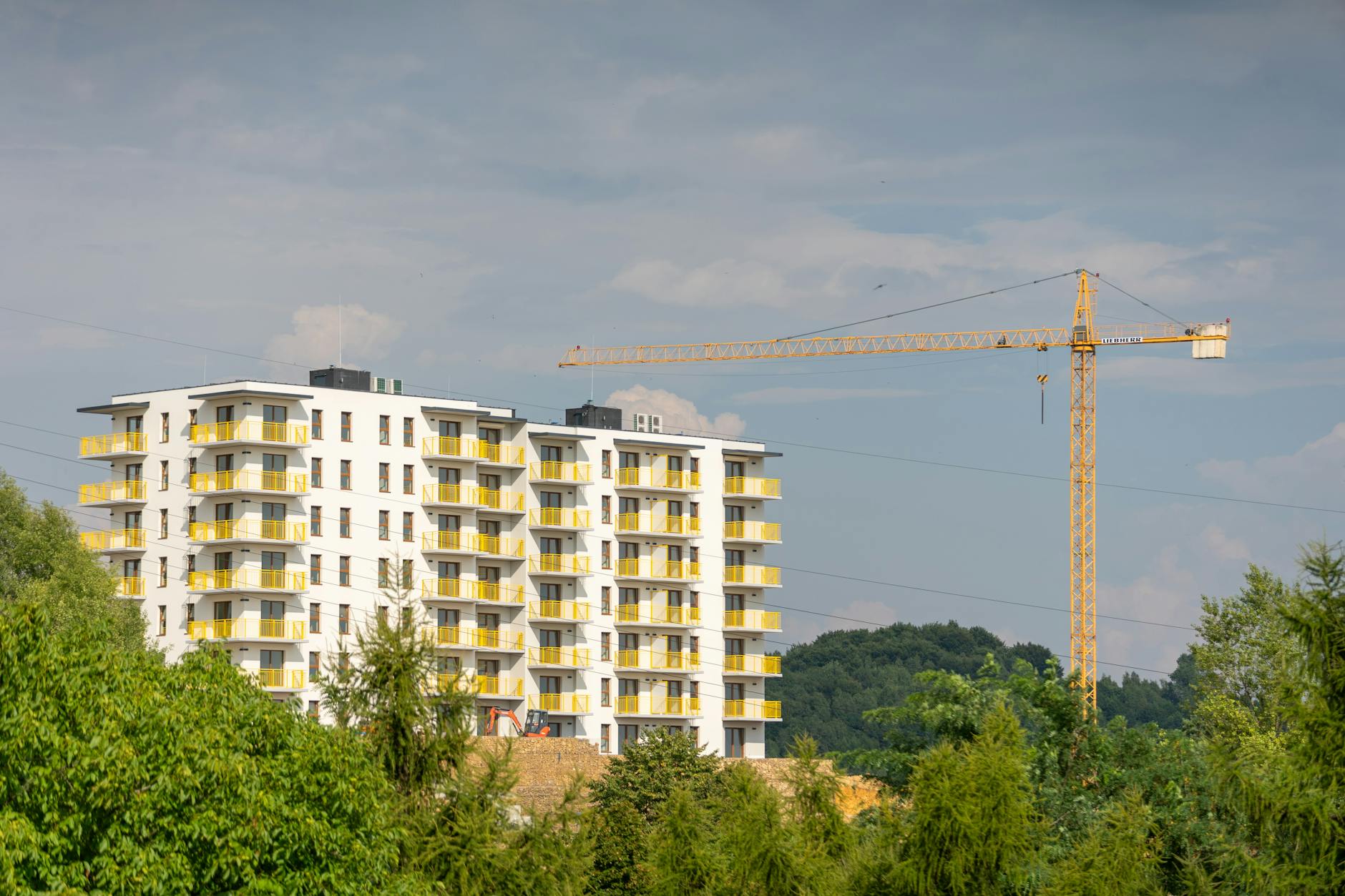 New multi-storey residential block with yellow balconies under construction beside a tall tower crane and green trees