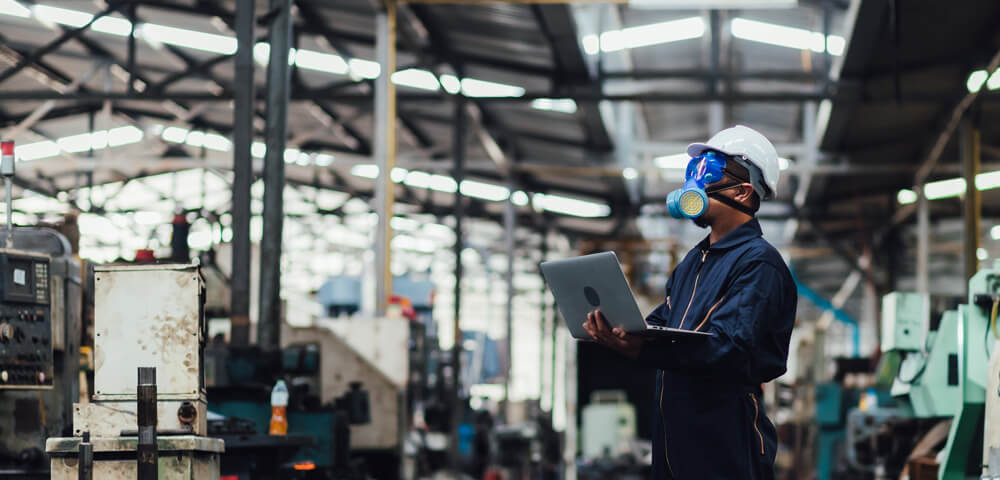 A young person with curly hair smiles at the camera while sitting in front of a laptop against a dark background.