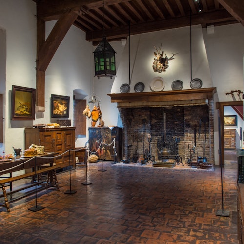 Et rustikt rum med en murstenspejs, træbjælker, malerier og antikke møbler under et højt loft.