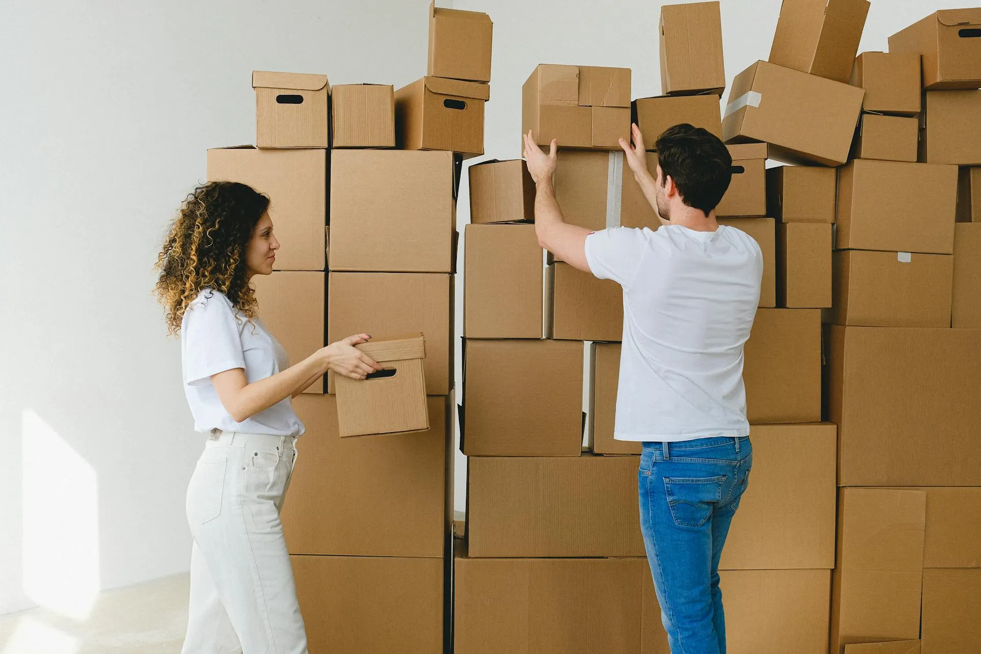 Young couple organizing and stacking numerous cardboard boxes together during a home or office relocation.