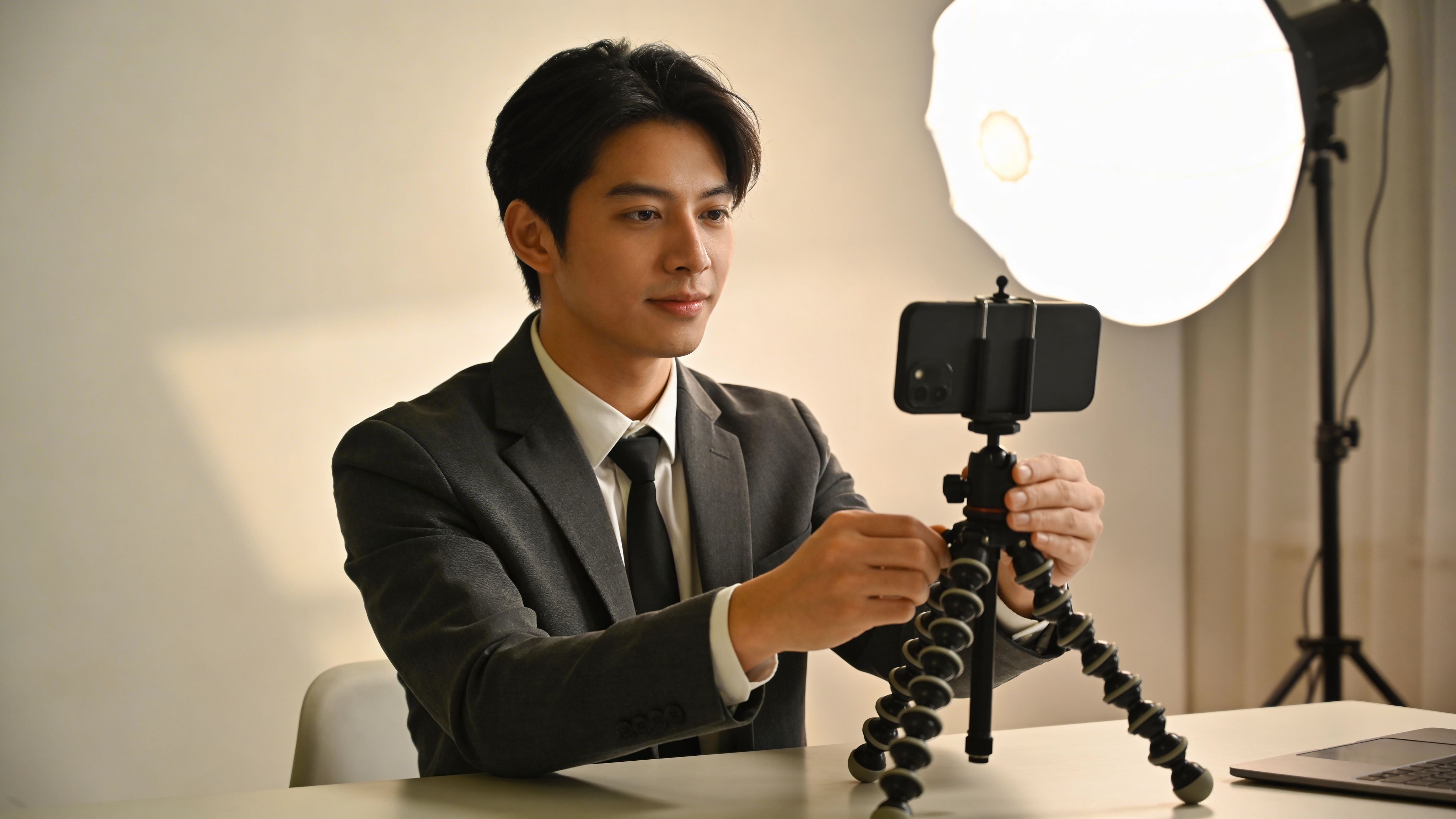 Ein junger Geschäftsmann in Anzug und Krawatte stellt sein Smartphone auf einem Stativ für eine Videokonferenz ein.