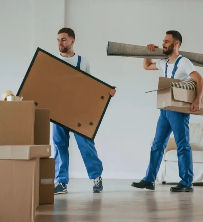 Professional movers carrying a large picture frame and a rolled rug in a new home.