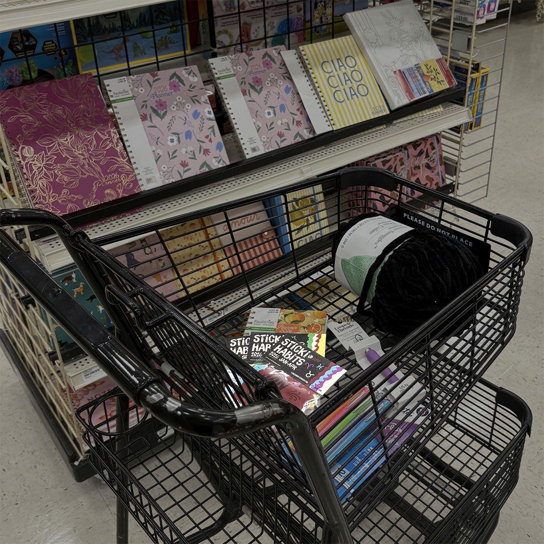 A shopping cart in a craft store aisle containing planner supplies, yarn, pens, and multiple packs of “Sticki Habits” zodiac calendar stickers displayed among other stationery products.