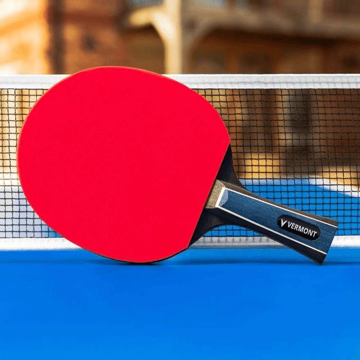 A red ping pong paddle rests on a blue table, with a blurred background suggesting an outdoor setting.