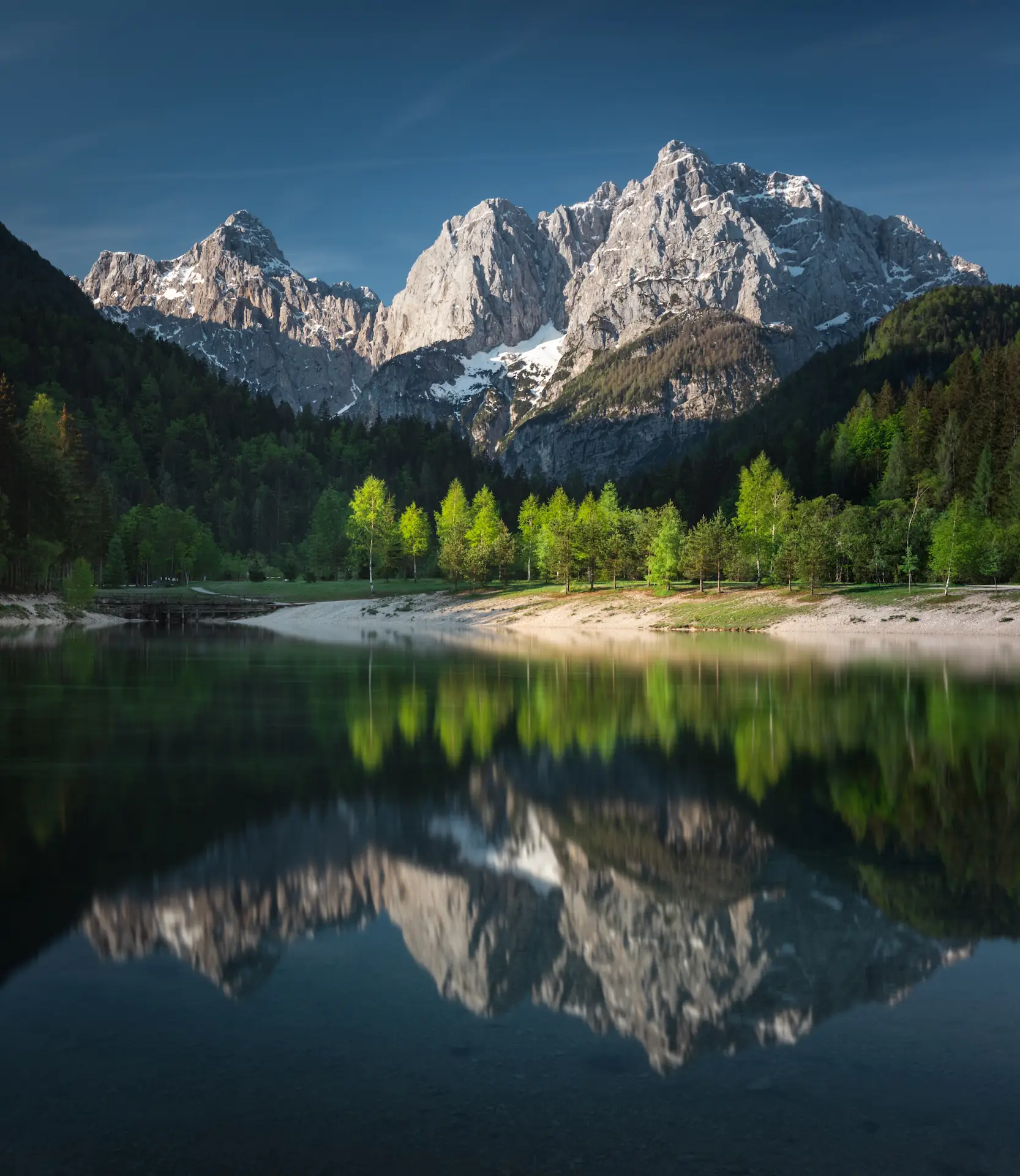 Snow-capped mountains reflect in tranquil lake Jasna, Slovenia, surrounded by lush greenery under a clear blue sky.