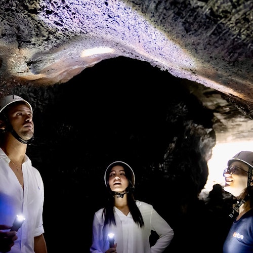 Três pessoas com capacetes e lanternas frontais exploram uma caverna pouco iluminada, usando lanternas de mão para iluminar o teto da caverna.
