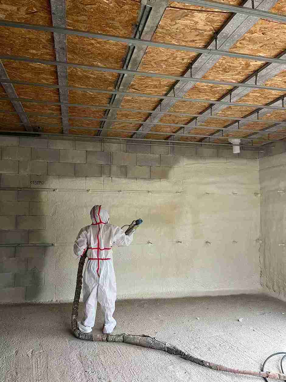 Une personne portant des équipements de protection pulvérise de la mousse polyuréthane projetée sur un mur en béton dans une pièce inachevée. Le plafond présente des structures métalliques et en bois, tandis que le sol reste en béton brut.