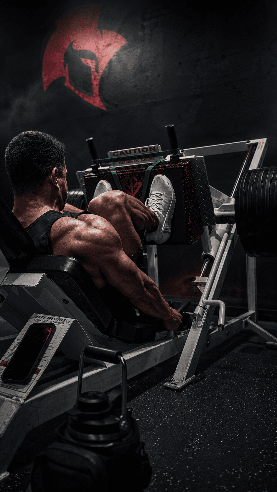 An athlete performs a focused leg press in a dim, high-contrast gym beneath a Spartan helmet emblem, embodying MORA's fusion of discipline and modern training.