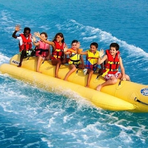 六个穿着救生衣的孩子在清澈的蓝色水面上骑着黄色的香蕉船，他们挥手并微笑着。