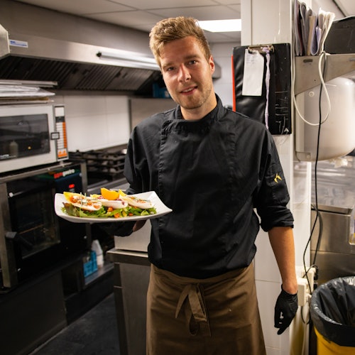 A chef in a black uniform holds a plated dish in a commercial kitchen.