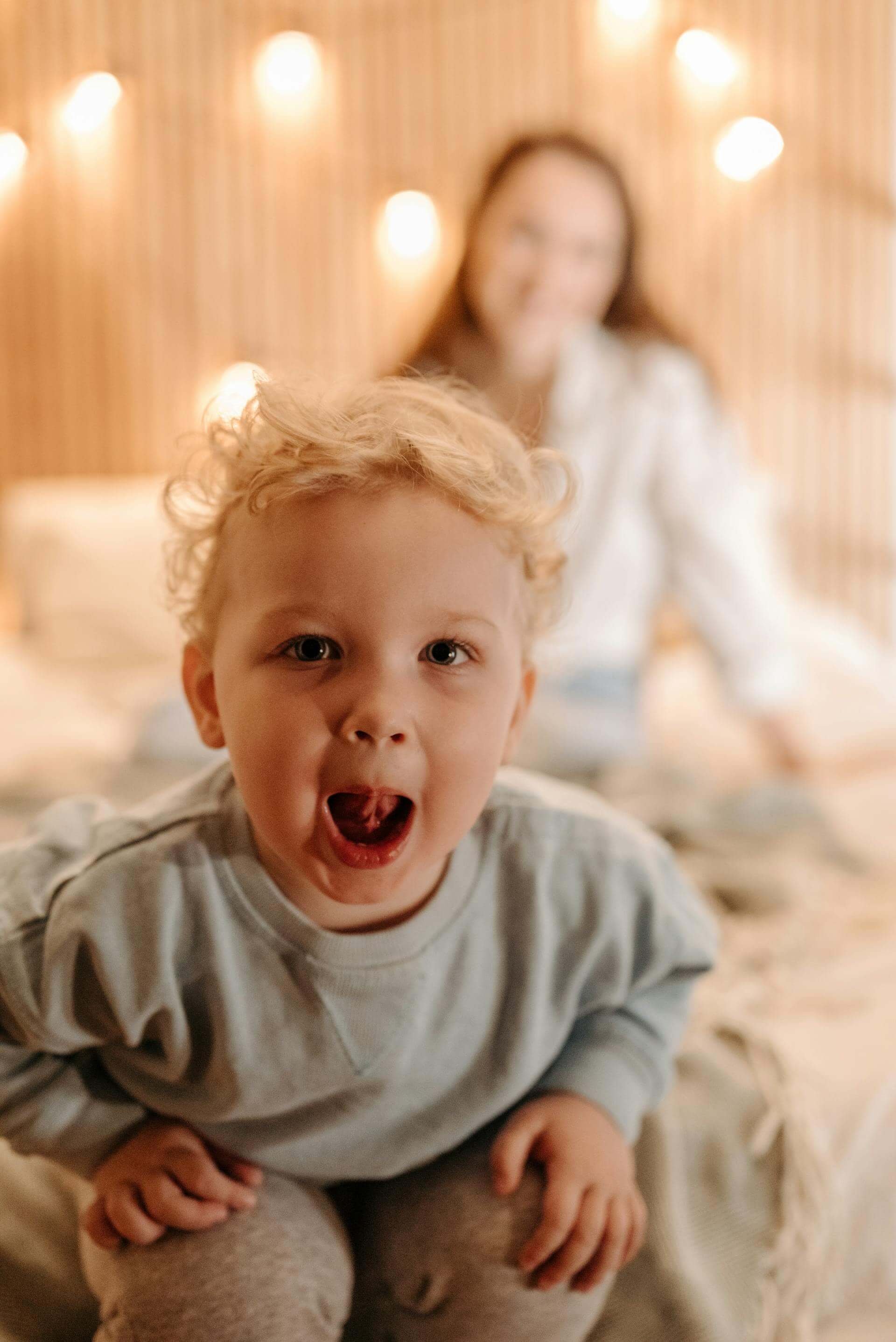 A little boy sits on a bed, with a woman softly visible in the background, creating a warm and cozy atmosphere