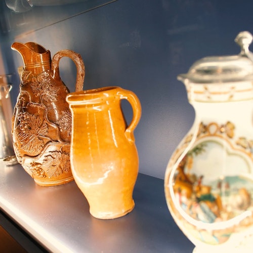 Tre keramikkaraffer visas på en hylla, en är intrikat snidad, en är slät orange, och en är målad med detaljerade mönster.