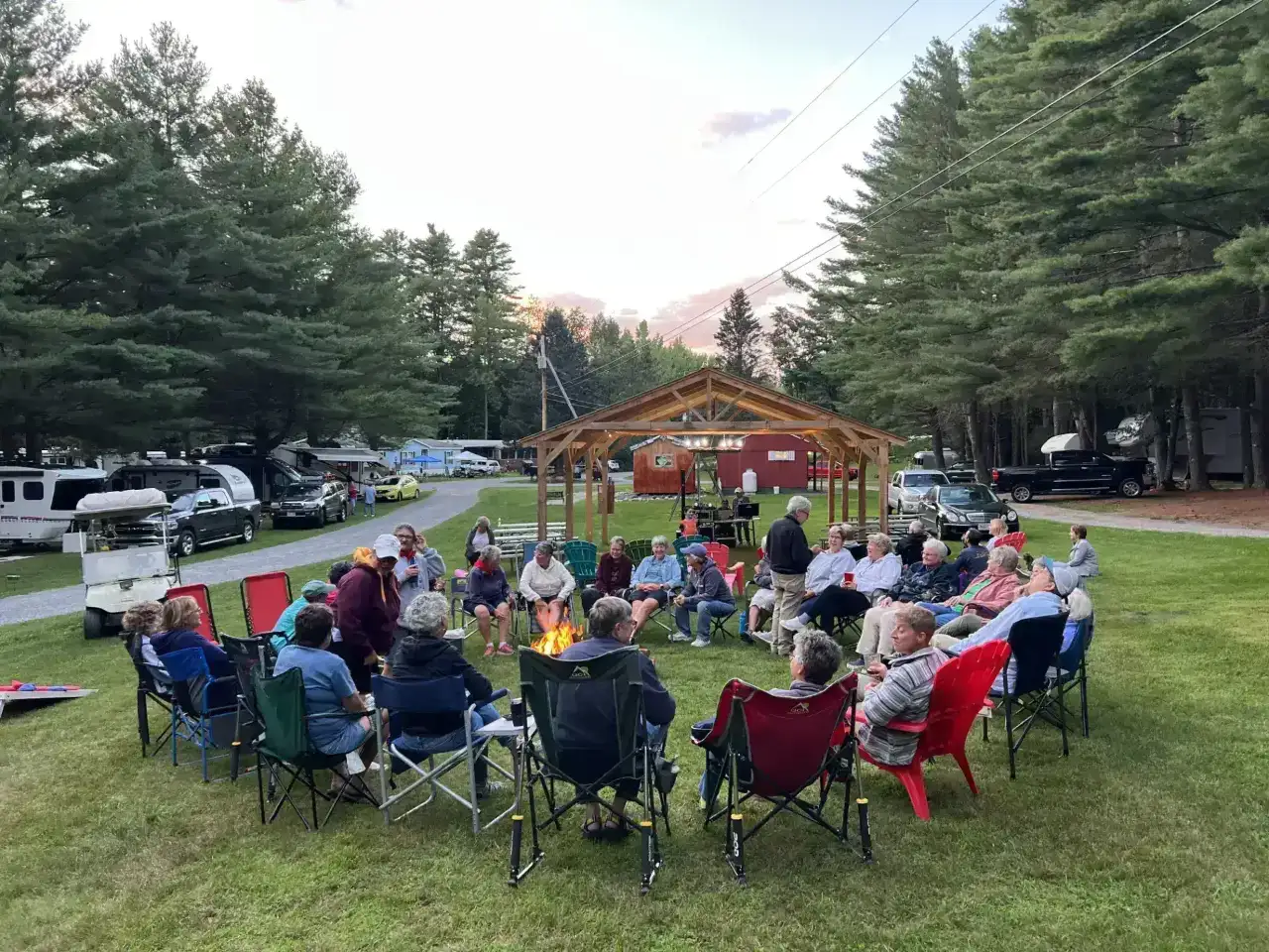 A large group of seasonal campers gathering in a circle around a community campfire at Pine Hollow Campground, a top-rated destination for southern Vermont campgrounds.