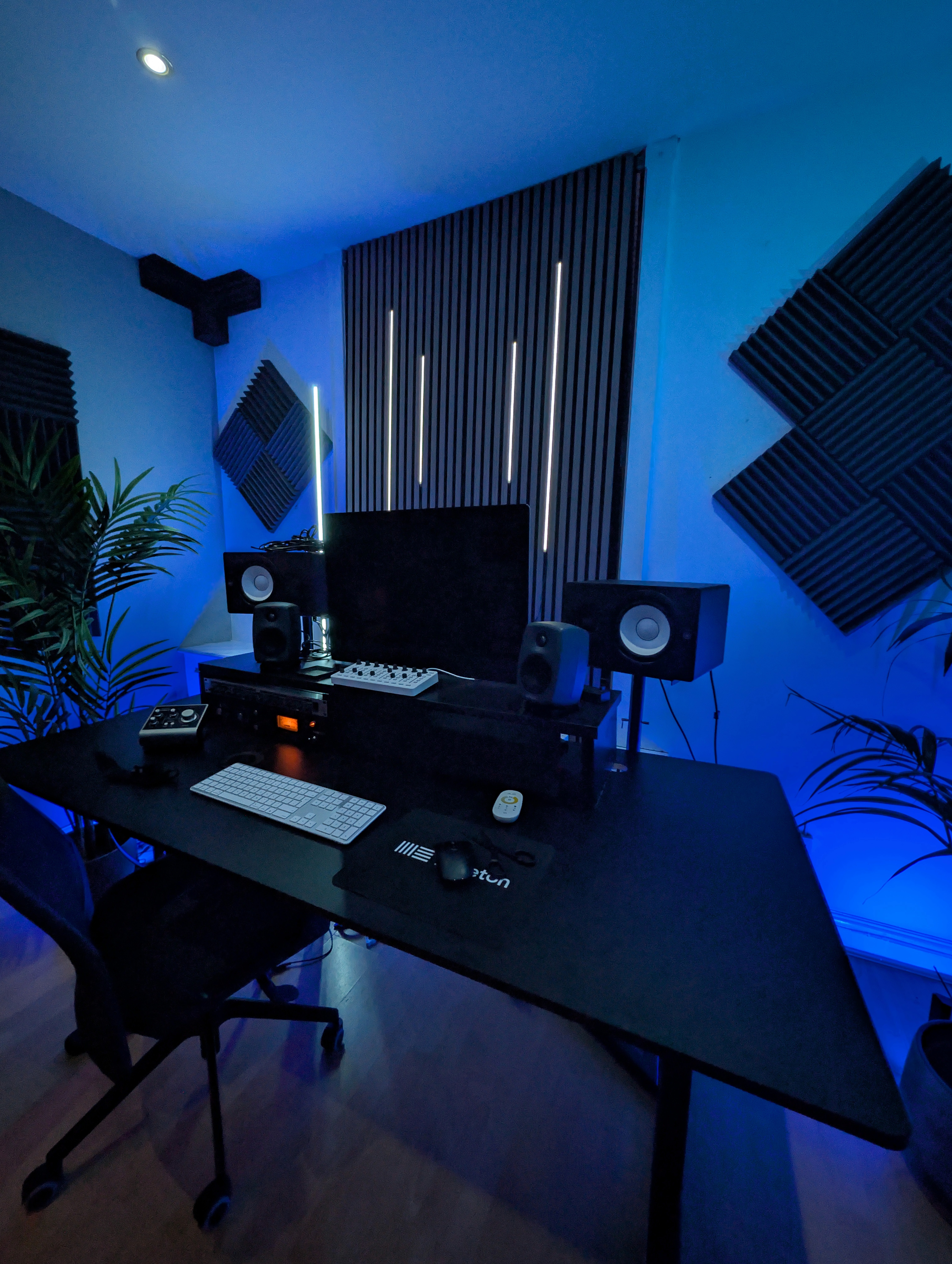 Modern music studio with a black desk, computer monitor, speakers, keyboard, and soundproofing panels. Blue lighting.