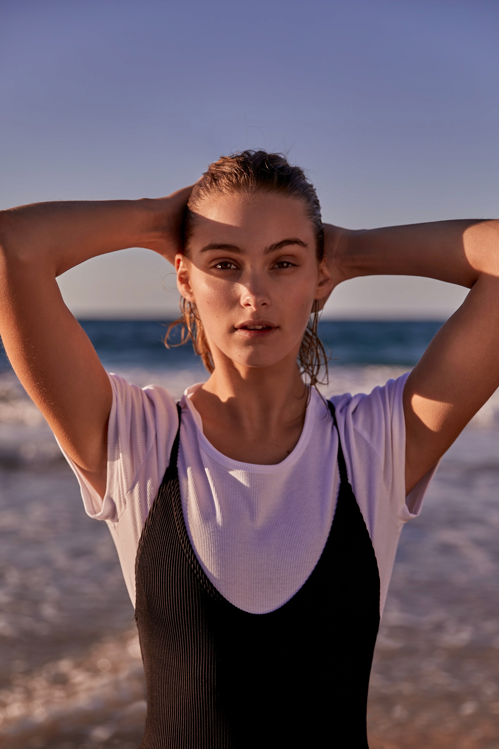 Woman in black swimsuit layered over sheer white t-shirt with hands behind head, photographed at the beach during golden hour with ocean in background.
