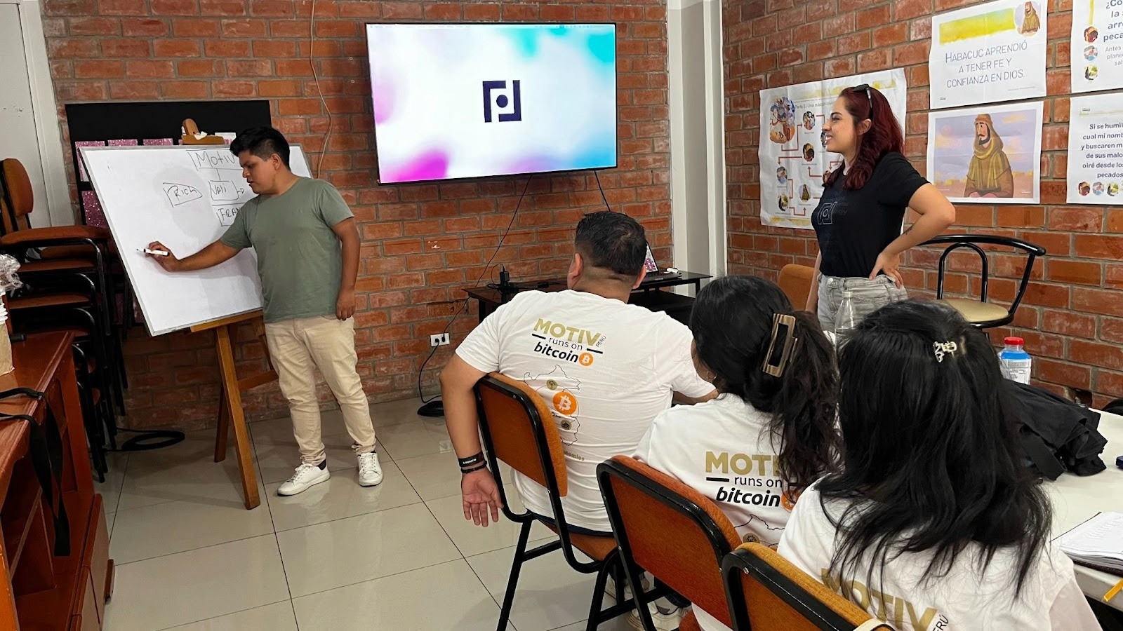 A Bitcoin education session hosted by Motiv in Peru. A facilitator writes on a whiteboard showing a diagram labelled "Motiv," while participants wearing "Motiv Runs on Bitcoin" t-shirts follow along. The Fedi logo is displayed on a screen at the front of the room.