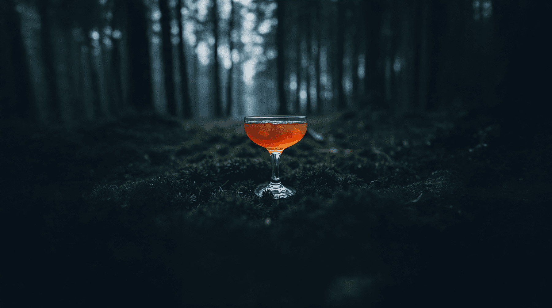 An amber cocktail in a glass sitting on some moss in a dark forest.