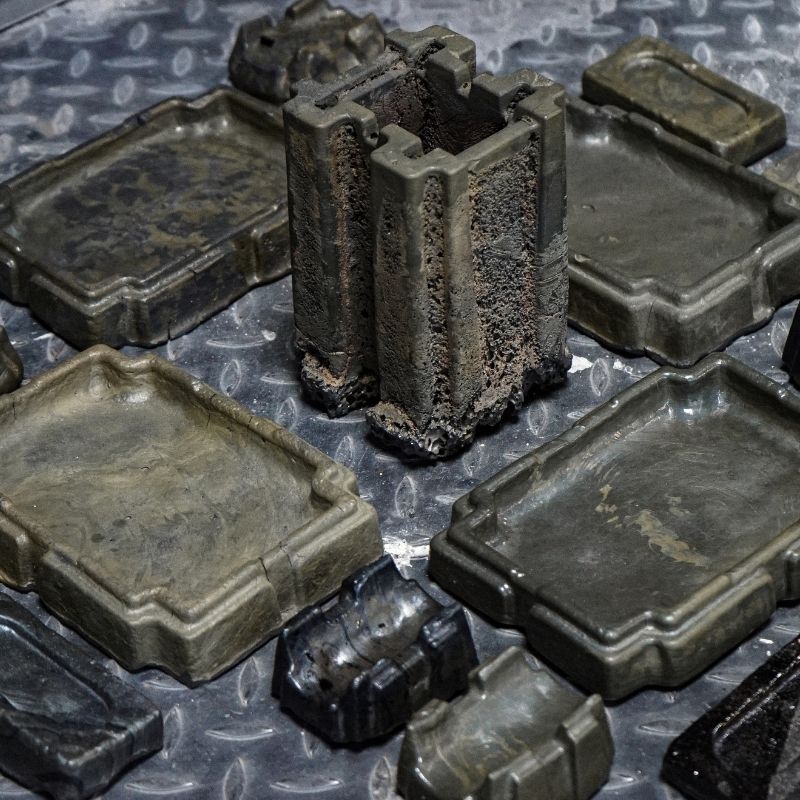 Lava objects designed by Max Greiner, including rectangular trays, small holders and a tall, textured vessel in dark grey and olive tones, displayed on metal flooring.
