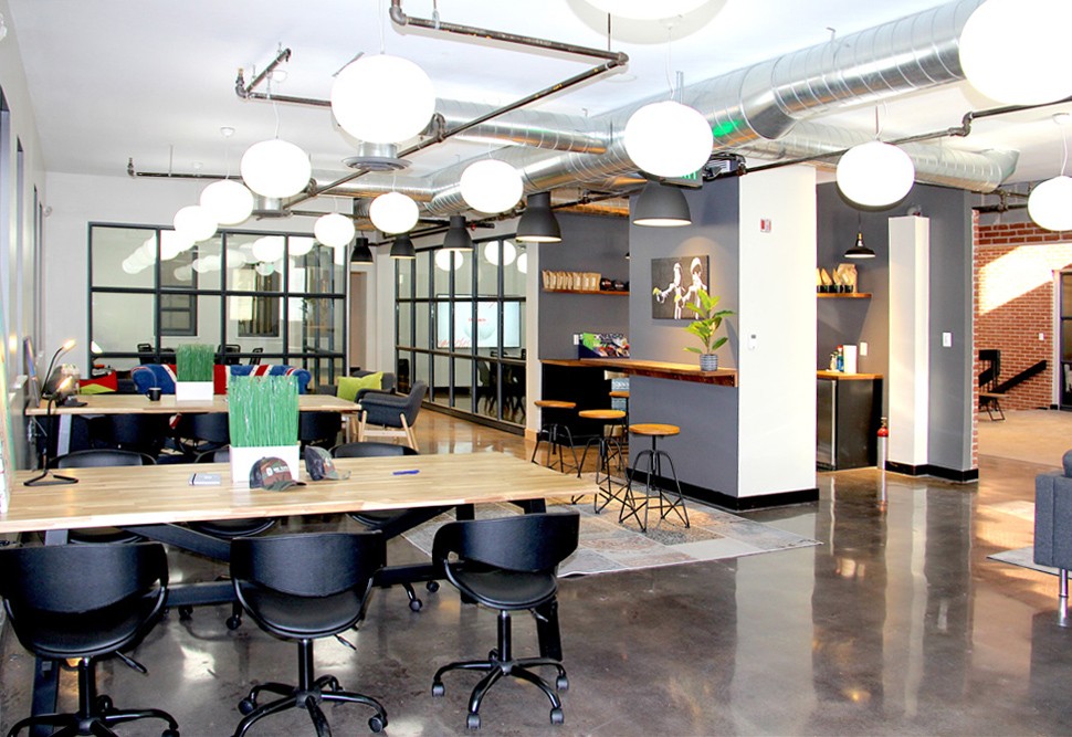 Modern coworking space with shared desks, chairs, and lounge areas, featuring industrial-style lighting and decor.