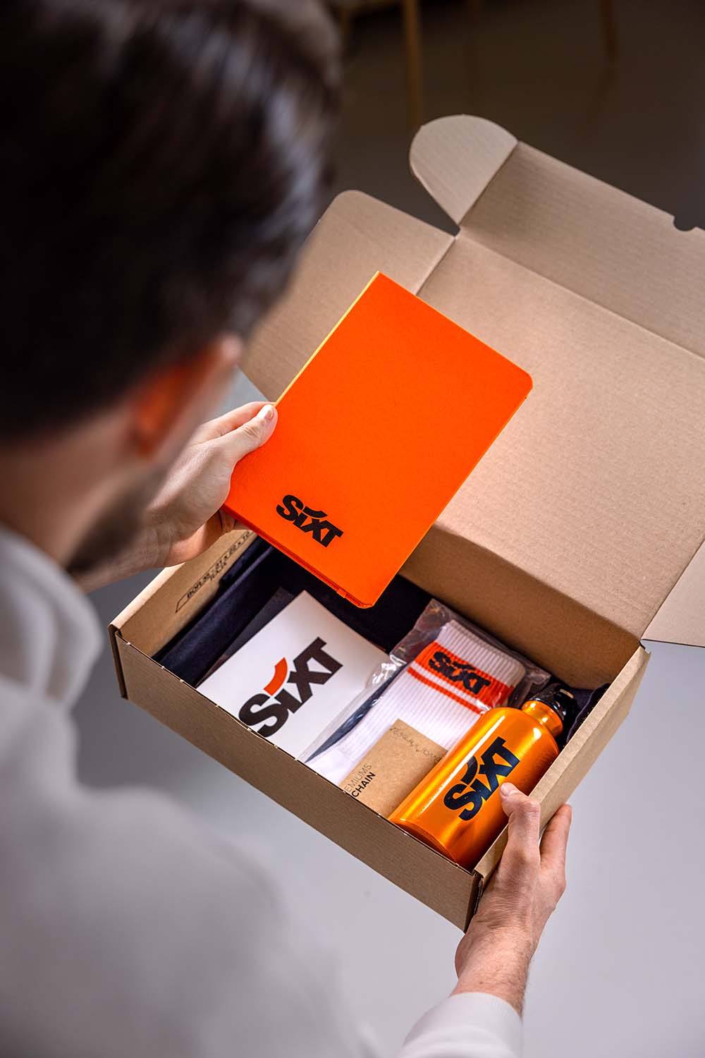 A person opens a cardboard box containing various items, including a bright orange box and other supplies.