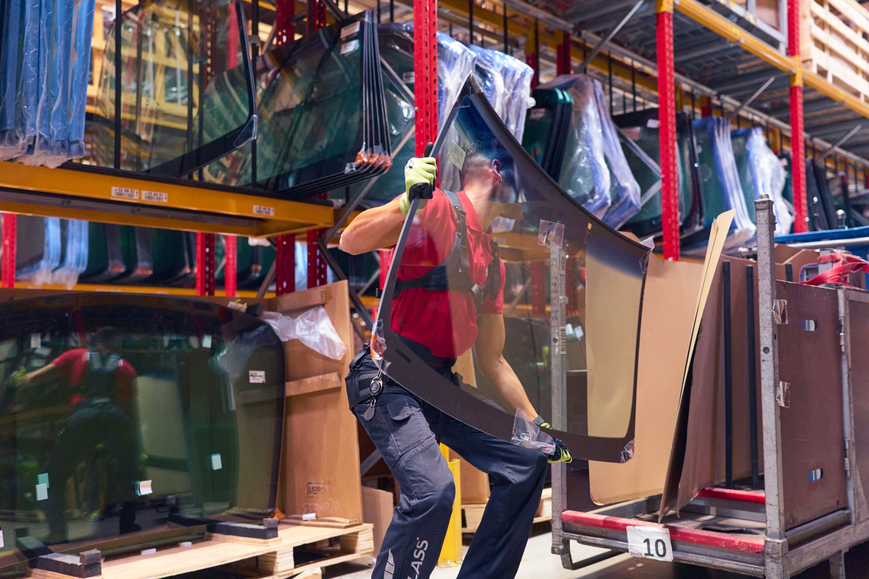 Travailleur en entrepôt soulevant une grande vitre de véhicule en portant l’exosquelette LiftSuit d’Auxivo, conçu pour réduire la charge lombaire lors de la manutention d’objets lourds et encombrants.
