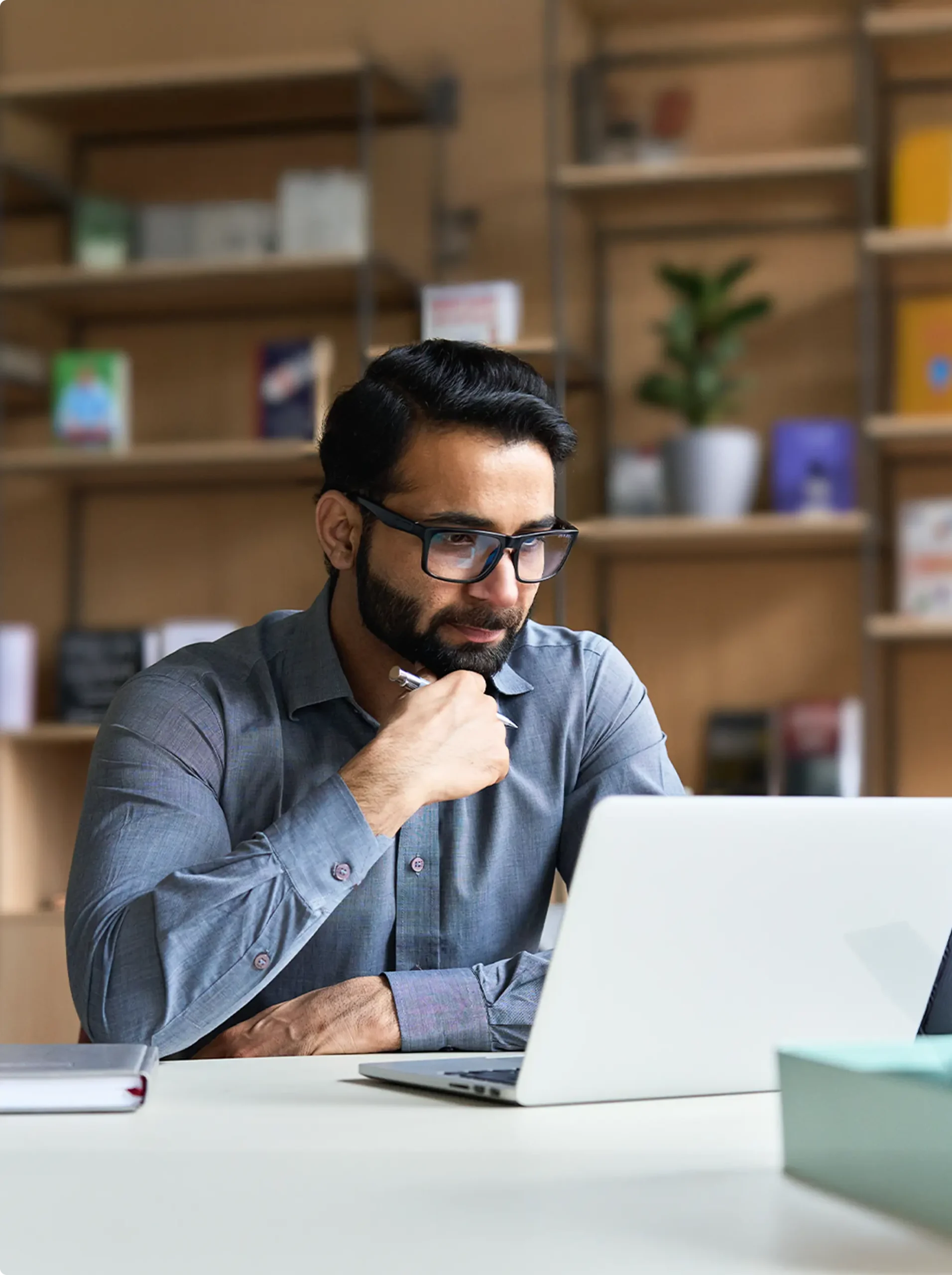 A man in a blue shirt sits thoughtfully at a desk, using a laptop in a well-lit, cozy workspace.