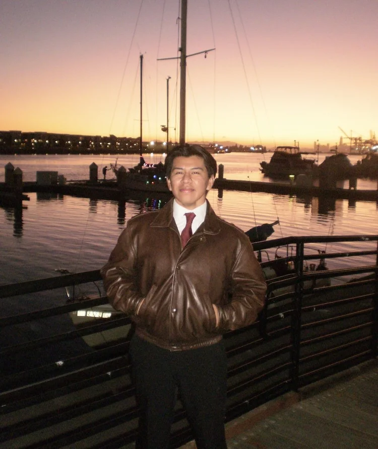 Uma pessoa está de pé em um píer ao pôr do sol, vestindo uma jaqueta de couro marrom e uma gravata vermelha, com veleiros e água calma na marina ao fundo, sob um vibrante céu laranja-rosado.